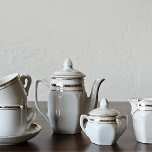 May include: A white porcelain tea set featuring a teapot with a lid, a lidded sugar bowl, a creamer, and two stacked teacups on a saucer. Each piece is decorated with a thin, horizontal gold band around the upper rim and body.