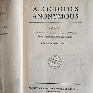 May include: Open book with the title "ALCOHOLICS ANONYMOUS" in large, bold, black letters. The text below reads "The Story of How Many Thousands of Men and Women Have Recovered from Alcoholism." A circular logo with "AA" is visible.