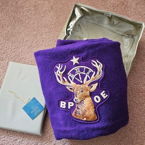 May include: A folded purple blanket with a large patch featuring a deer head, clock, and star. The blanket is next to a gray package with a blue tag and a clear storage bag.