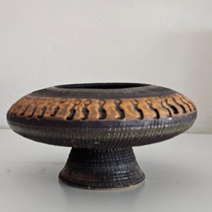 May include: A ceramic pedestal bowl with a wide, flat rim and a textured, dark brown base. The rim features a band of tan-colored cut-out designs. The bowl's interior is black, and the overall shape is reminiscent of a mushroom.