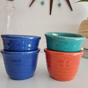 May include: Four small, ceramic bowls in shades of blue, teal, and orange are stacked in pairs. The bowls have a rounded shape with a slightly flared rim. The glossy finish reflects light, highlighting the vibrant colors. These bowls are suitable for serving dips or small portions.