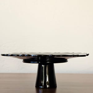May include: A glossy black glass cake stand featuring a wide, circular plate with a scalloped, decorative edge. The stand is supported by a sturdy, tapered pedestal base, providing a raised platform for display purposes.