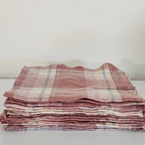 May include: A stack of pink and white plaid fabric squares. The fabric has a soft, textured appearance with a light pink base and a plaid pattern in white and light blue. The edges of the squares are slightly uneven.