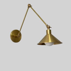 May include: A brass wall-mounted lamp with an adjustable arm and a conical shade. The lamp features a round base and a pivoting arm that extends to the shade. The shade houses a white light bulb. The fixture is made of brass.