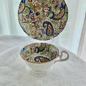 May include: A white teacup and saucer set with a vibrant floral and paisley design. The teacup features a gold rim and handle. The round saucer has a matching pattern. The set is presented on a clear stand.