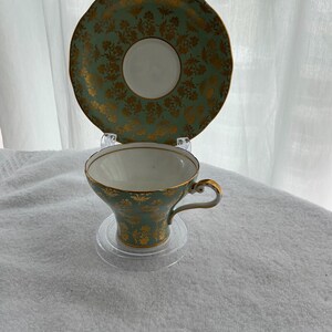 May include: A vintage teacup and saucer set. The cup and saucer are a pale green with a gold floral pattern. The cup has a gold handle and trim. The saucer has a scalloped edge. The set is displayed on a clear plastic stand.