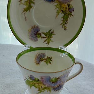 May include: A white porcelain teacup and saucer set featuring a painted thistle pattern in shades of purple and green. The rim of both pieces is accented with a thin green band and gold trim. The cup has a gold base and a single curved handle.