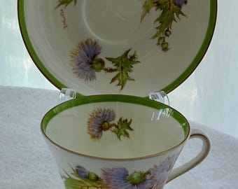 May include: A white porcelain teacup and saucer set featuring a painted thistle pattern in shades of purple and green. The rim of both pieces is accented with a thin green band and gold trim. The cup has a gold base and a single curved handle.