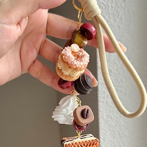 May include: A whimsical keychain featuring miniature food charms. The charms include a pink frosted donut, a chocolate cookie, a white cream swirl, and a biscuit with the word "Lotus" on it. The keychain is attached to a beige cord.