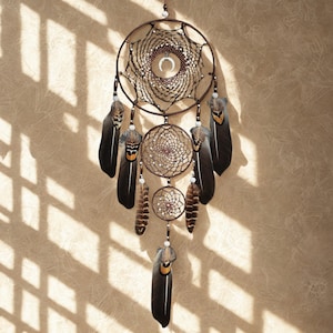May include: A handcrafted dreamcatcher with a large circular frame and multiple smaller circular designs. The dreamcatcher features a web-like pattern, beads, and dark feathers with orange and yellow accents. It hangs against a neutral-toned background.