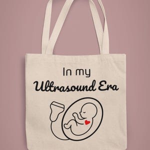Op de afbeelding: Beige draagtas met de tekst "In my Ultrasound Era" en een zwarte lijntekening van een foetus in een ovaal met een rood hart. De tas heeft twee crèmekleurige handvatten.
