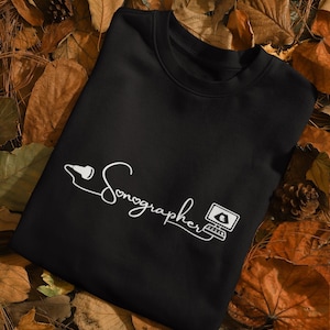 May include: Black sweatshirt with the word "Sonographer" in a white script font, accompanied by a stylized sonogram image and a computer monitor graphic. The sweatshirt is displayed on a bed of autumn leaves.