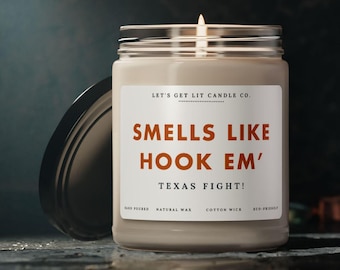 May include: A white candle with a black lid. The candle has a label that reads "Let's Get Lit Candle Co." and "Smells Like Hook 'Em' Texas Fight!" The label also includes the text "Hand Poured, Natural Wax, Cotton Wick, Eco-Friendly."