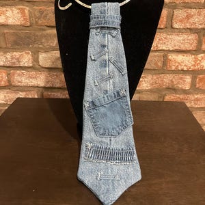 Puede incluir: Una corbata de mezclilla única con un pequeño bolsillo y otros detalles de mezclilla. La corbata se exhibe en una cabeza de maniquí negra con un collar plateado. El fondo es una pared de ladrillos.