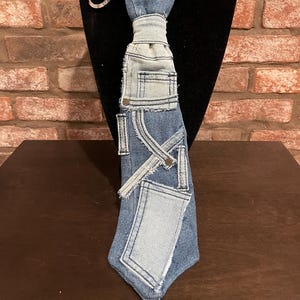 Puede incluir: Una corbata de mezclilla con parches y costuras en azul claro y azul medio. La corbata se exhibe en un maniquí negro con un collar plateado. La corbata tiene un diseño único, con la apariencia de bolsillos y costuras de jeans.