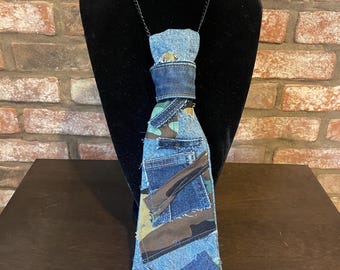 May include: A handmade denim tie with a camouflage pattern. The tie is constructed from various denim patches and features a dark blue denim band around the neck. It is displayed on a black mannequin head.