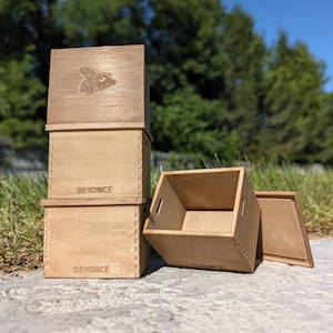 Puede incluir: Una pila de dos cajas de madera de color marrón claro con esquinas de unión dentada y el nombre BEYONCE grabado en el frente. Una tercera caja de madera abierta se encuentra junto a la pila con su tapa a juego colocada plana en el suelo.