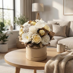 Peut inclure: Un bouquet de fleurs blanches, crème et jaunes, avec des chocolats dorés, est présenté dans une boîte ronde beige. L'arrangement est posé sur une table en bois, avec une couverture et un mug beiges. En arrière-plan, une fenêtre et un canapé.