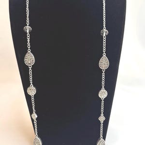 May include: A silver-toned necklace with a chain design. The necklace features teardrop and round pendants with a textured, sparkly surface, interspersed with clear, faceted beads. The necklace is displayed on a black mannequin.