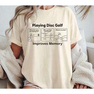 Puede incluir: Camiseta color crema con el texto "Playing Disc Golf Improves Memory". La camiseta presenta un gráfico humorístico de figuras de palo que representan las frustraciones comunes del disc golf. Es una prenda cómoda e informal.