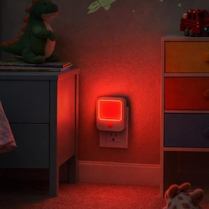 Puede incluir: Una luz de noche roja enchufada en un enchufe de pared en el dormitorio de un niño. La habitación tiene una cama azul, una mesita de noche con un dinosaurio de peluche y una cómoda colorida. La pared tiene estrellas y un cohete que brillan en la oscuridad.