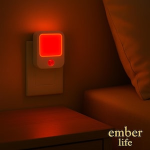 May include: A white night light with a red square light is plugged into a wall outlet. The night light has a small red button. The text "ember life" is in the bottom right corner. The night light is on a wall next to a wooden nightstand.