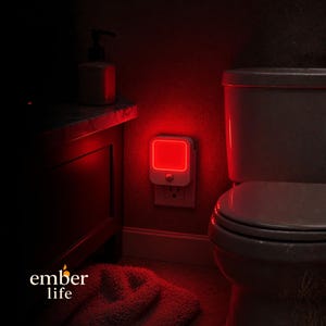 Puede incluir: Una luz de noche blanca con un cuadrado rojo brillante está enchufada a una toma de corriente en una pared roja. La luz de noche está en un baño, con un inodoro y un armario visibles. El texto "ember life" está en la parte inferior.