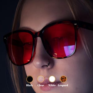 May include: Square-framed sunglasses with red lenses and a tortoiseshell pattern. The image shows the sunglasses on a person, with options for black, clear, white, and leopard frames. The text below the options reads "Black", "Clear", "White", and "Leopard".