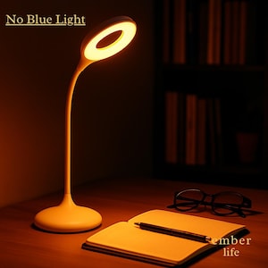 May include: A modern, cream-colored desk lamp with a circular light head, illuminating a warm glow. The lamp is on a wooden desk with a notebook, pen, and glasses. The text "No Blue Light" is visible.