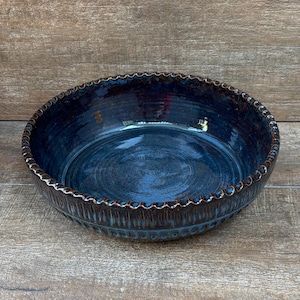 May include: A handmade, round ceramic bowl with a deep blue glaze. The rim features a textured, scalloped design with a dark brown edge. The bowl's interior has a glossy finish, reflecting light. This decorative bowl is suitable for serving or display.