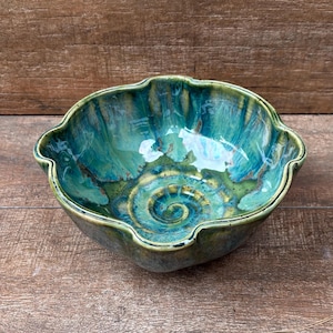 May include: A ceramic bowl with a scalloped rim and a glossy glaze featuring swirling patterns of deep teal, turquoise, and olive green. The interior base displays a prominent spiral design, while the exterior maintains a smooth, rounded shape.