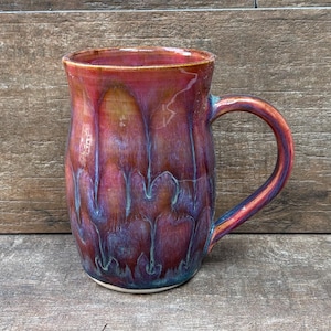 May include: A ceramic mug featuring a glossy glaze with a blend of deep red, magenta, and purple tones. The surface displays a vertical drip pattern with subtle blue and violet highlights, a wide cylindrical body, and a curved handle on the side.