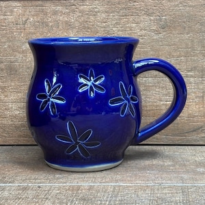 May include: A cobalt blue ceramic mug with a rounded body and a large handle. The mug features a floral design with white outlines. The mug is sitting on a wooden surface.