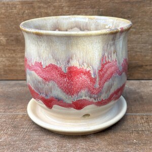 May include: A ceramic planter with a rounded shape and a matching saucer. The glaze features a creamy beige base color with a prominent, horizontal, wavy band of vibrant red that drips down the sides, creating a layered, textured appearance.