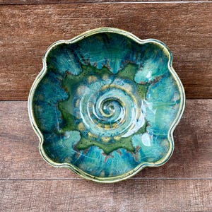 May include: A scalloped ceramic bowl featuring a glossy glaze in shades of teal, turquoise, and forest green. The interior surface displays a prominent spiral pattern radiating from the center, with blended color transitions and dark speckles.