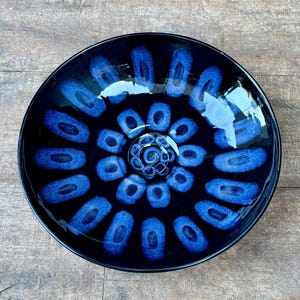 May include: A round ceramic bowl with a glossy black glaze featuring a hand-painted pattern of vibrant blue oval shapes arranged in concentric circles radiating from a central spiral design at the base of the interior of the dish.