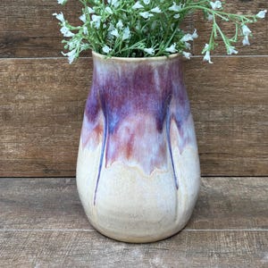 May include: A ceramic vase with a rounded body and a tapered neck, featuring a glaze that transitions from a deep purple and blue at the top to a creamy off-white at the base. The vase is filled with a cluster of small white artificial flowers.