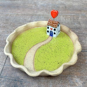May include: A round ceramic trinket dish with a scalloped edge, featuring a speckled light green interior and a tan path leading to a small white house sculpture. The house has a brown roof, a blue door, and a red heart topper on a wire.