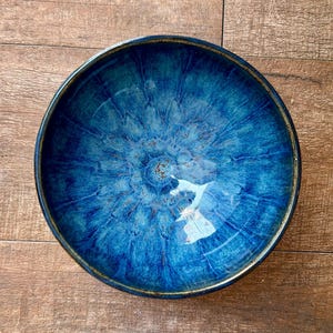 May include: A round, glazed ceramic bowl with a deep blue interior and a dark brown rim. The bowl's interior has a textured, swirling pattern with lighter blue and brown accents. The bowl is set against a wooden surface.