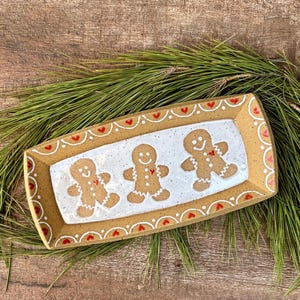 May include: A rectangular ceramic tray with a speckled tan border and a white center. The tray features three brown gingerbread figures with white icing details and small red hearts. The border is decorated with a white scalloped line and red hearts.