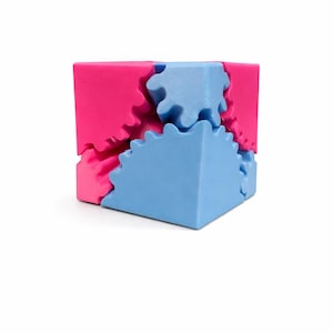 May include: A three-dimensional puzzle cube with interlocking gear-shaped pieces. The cube is composed of pink and blue sections, creating a visually engaging geometric design. The pieces fit together to form a complete cube.