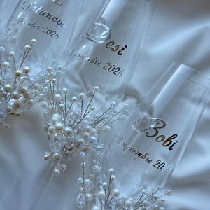 Puede incluir: Copas de champ&aacute;n transparentes con letras plateadas y adornos de perlas y cristales. Las copas est&aacute;n personalizadas con nombres y fechas, perfectas para una boda. Sobre un fondo blanco.