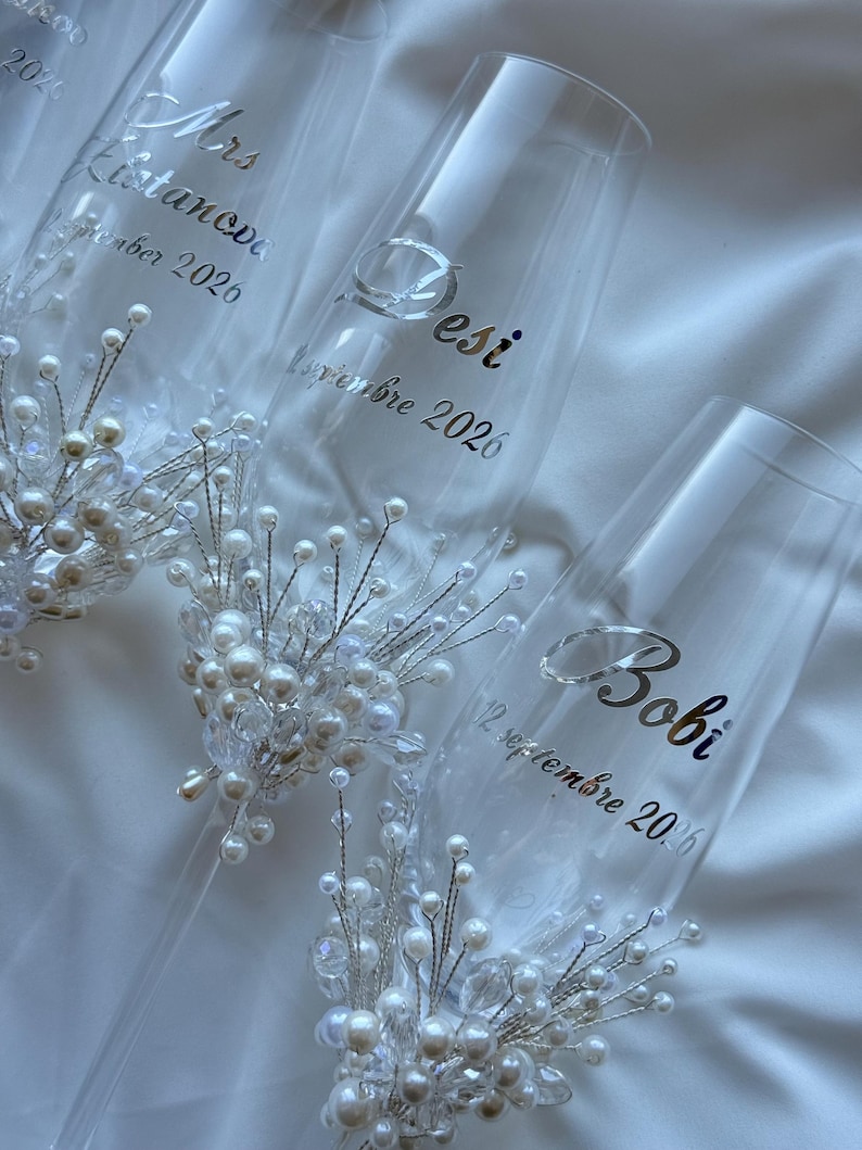 Puede incluir: Copas de champ&aacute;n transparentes con letras plateadas y adornos de perlas y cristales. Las copas est&aacute;n personalizadas con nombres y fechas, perfectas para una boda. Sobre un fondo blanco.