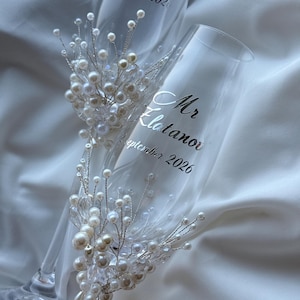 Puede incluir: Dos copas de champ&aacute;n transparentes con texto plateado "Mr. Elatanova" y "Mrs. Elatanova" con fecha. Cada copa est&aacute; adornada con un racimo de perlas blancas y cristales transparentes sobre alambre plateado, tema de boda.