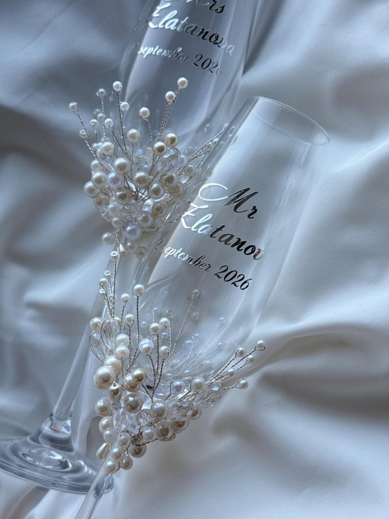 Puede incluir: Dos copas de champ&aacute;n transparentes con texto plateado "Mr. Elatanova" y "Mrs. Elatanova" con fecha. Cada copa est&aacute; adornada con un racimo de perlas blancas y cristales transparentes sobre alambre plateado, tema de boda.