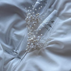 Puede incluir: Dos copas de champ&aacute;n transparentes con texto plateado "Mrs. Elatanos, 12 septiembre 2026". Cada copa est&aacute; adornada con perlas y cristales. Las copas descansan sobre una tela blanca y sedosa.