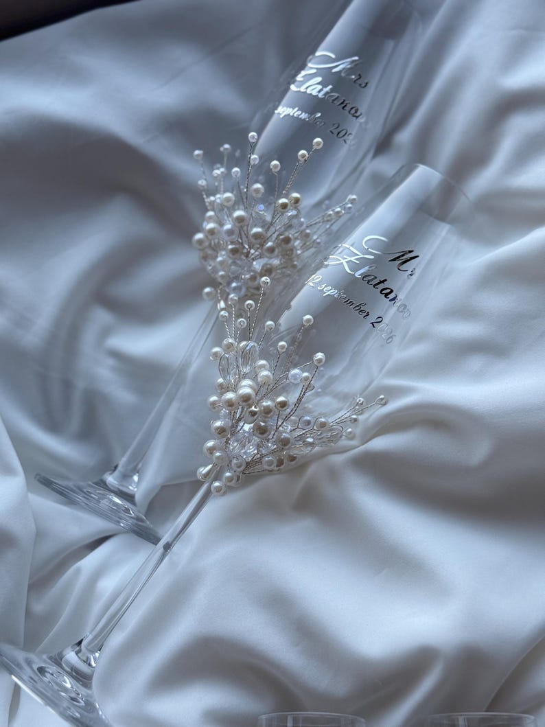 Puede incluir: Dos copas de champ&aacute;n transparentes con texto plateado "Mrs. Elatanos, 12 septiembre 2026". Cada copa est&aacute; adornada con perlas y cristales. Las copas descansan sobre una tela blanca y sedosa.