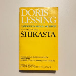 May include: A vintage paperback book titled "Shikasta" by Doris Lessing. The cover is bright yellow with white and black text. The book is part of the "Canopus in Argos: Archives" series and includes the text "Personal Psychological Historical Documents".