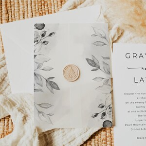 Puede incluir: Un conjunto de invitaciones de boda con una tarjeta translúcida con hojas grises de acuarela y un sello de cera beige. La invitación está dentro de un sobre blanco, con una tarjeta a juego visible a la derecha.