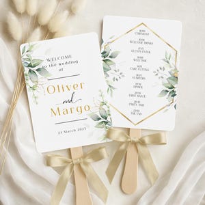 Könnte beinhalten: Zwei weiße Hochzeitsfächer mit goldenem Text und botanischen Akzenten. Auf einem Fächer steht "Willkommen zur Hochzeit von Oliver und Margo, 24. März 2023." Der andere Fächer zeigt den Hochzeitsablauf. Jeder Fächer hat einen Holzgriff und ein goldenes Band.